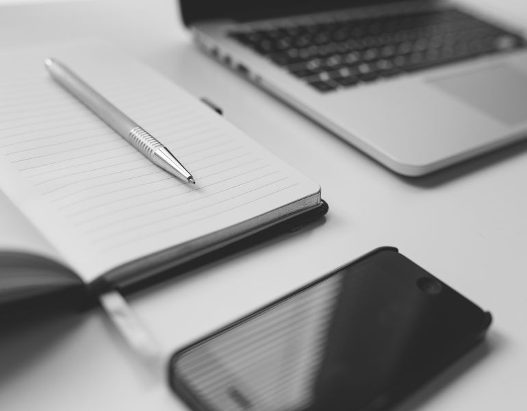 Schwarz-weiß Foto eines Notizbuchs, eines Stifts und eines Handys auf einem Schreibtisch.