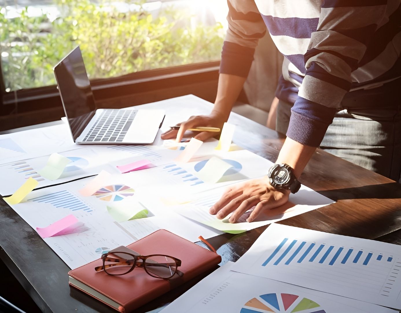 Geschäftsmann arbeitet an seinem Laptop am Schreibtisch, umgeben von Unterlagen und Diagrammen.